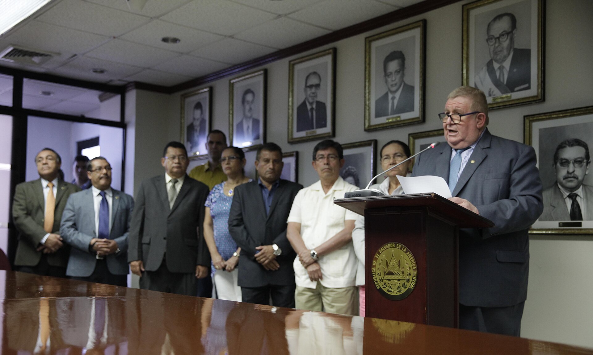 A man in a suit speaking at a lectern bearing the Salvadoran coat of arms, with officials standing behind him in front of framed portraits