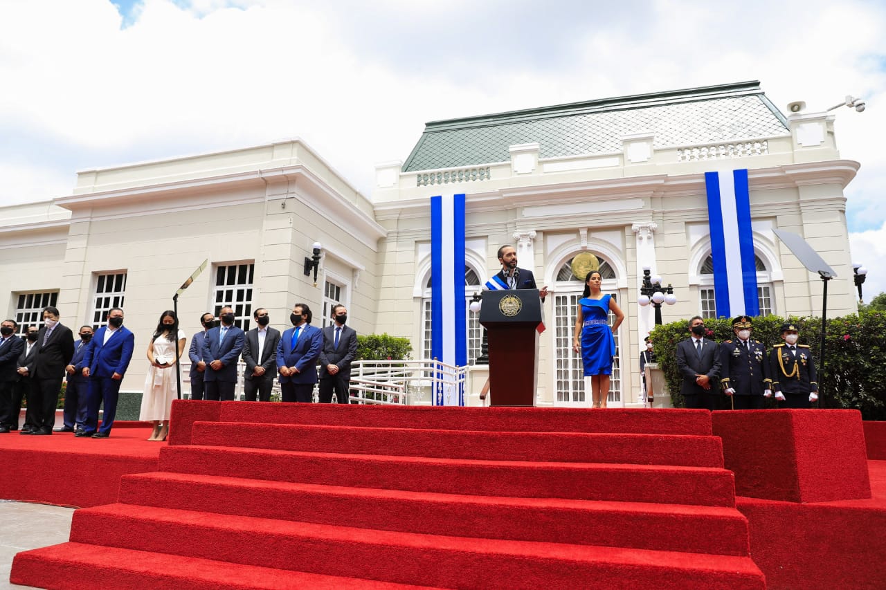 A president speaks at a podium on red-carpeted steps in front of the white Presidential House in San Salvador, with officials and honour guards standing by and blue-and-white flags hanging from the facade