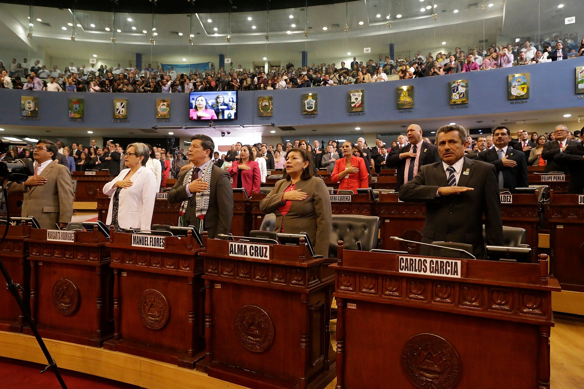 Deputies standing with hands on their chests at wooden desks in the debating chamber of El Salvador's Legislative Assembly, with a packed gallery behind