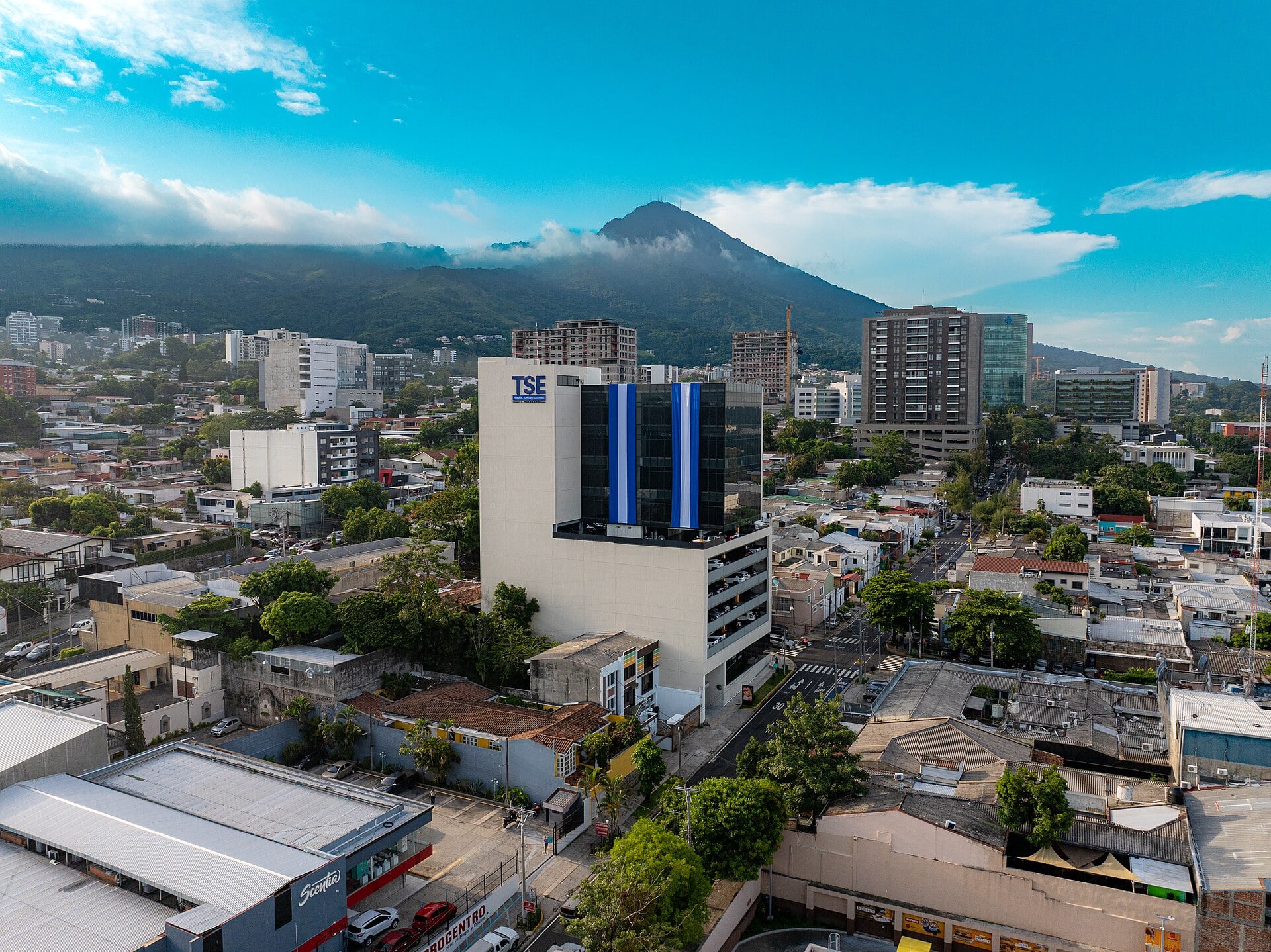 Aerial view of the Supreme Electoral Tribunal building in San Salvador with blue-and-white flag banners on its facade and a volcano behind