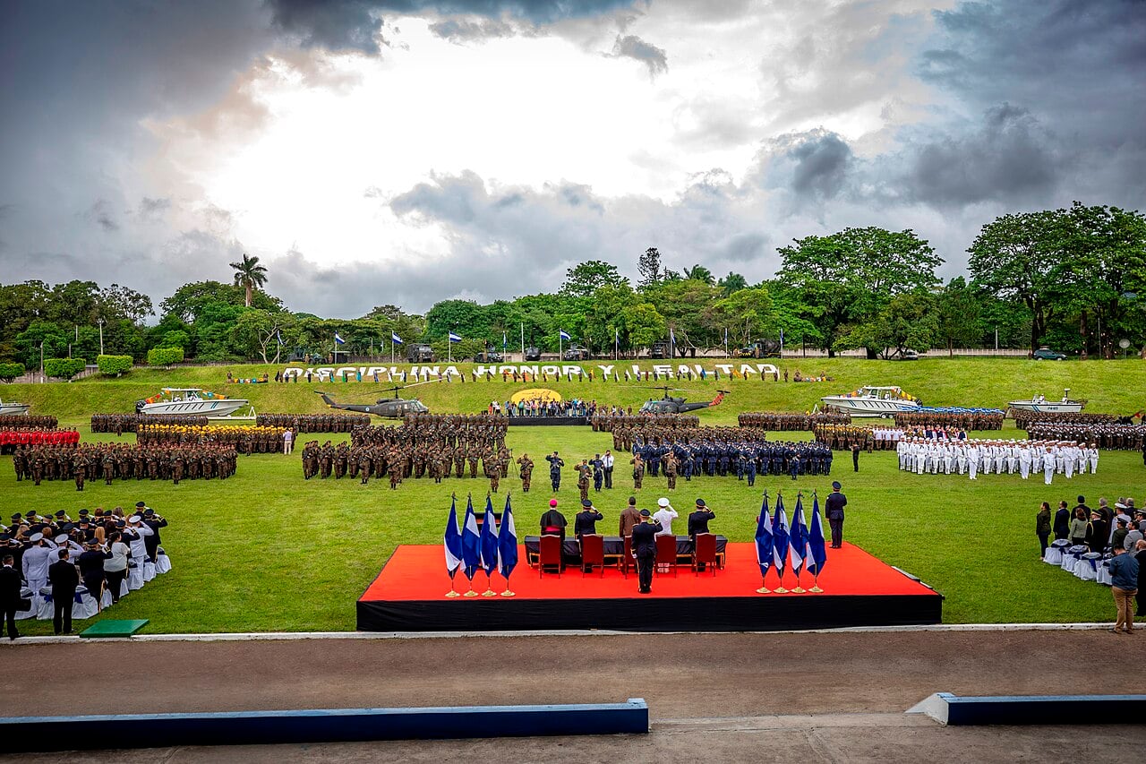 Soldiers, sailors and officers in formation on a green field at an armed forces ceremony in El Salvador, with helicopters and boats behind