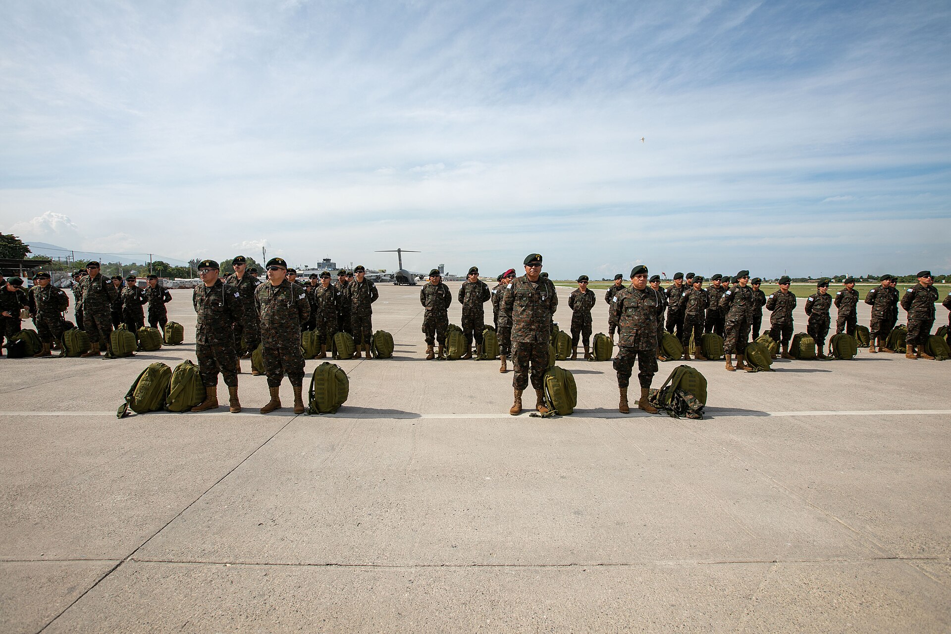 Salvadoran and Guatemalan troops stand in formation with backpacks on the airport apron at Port-au-Prince, Haiti, with a transport aircraft behind them
