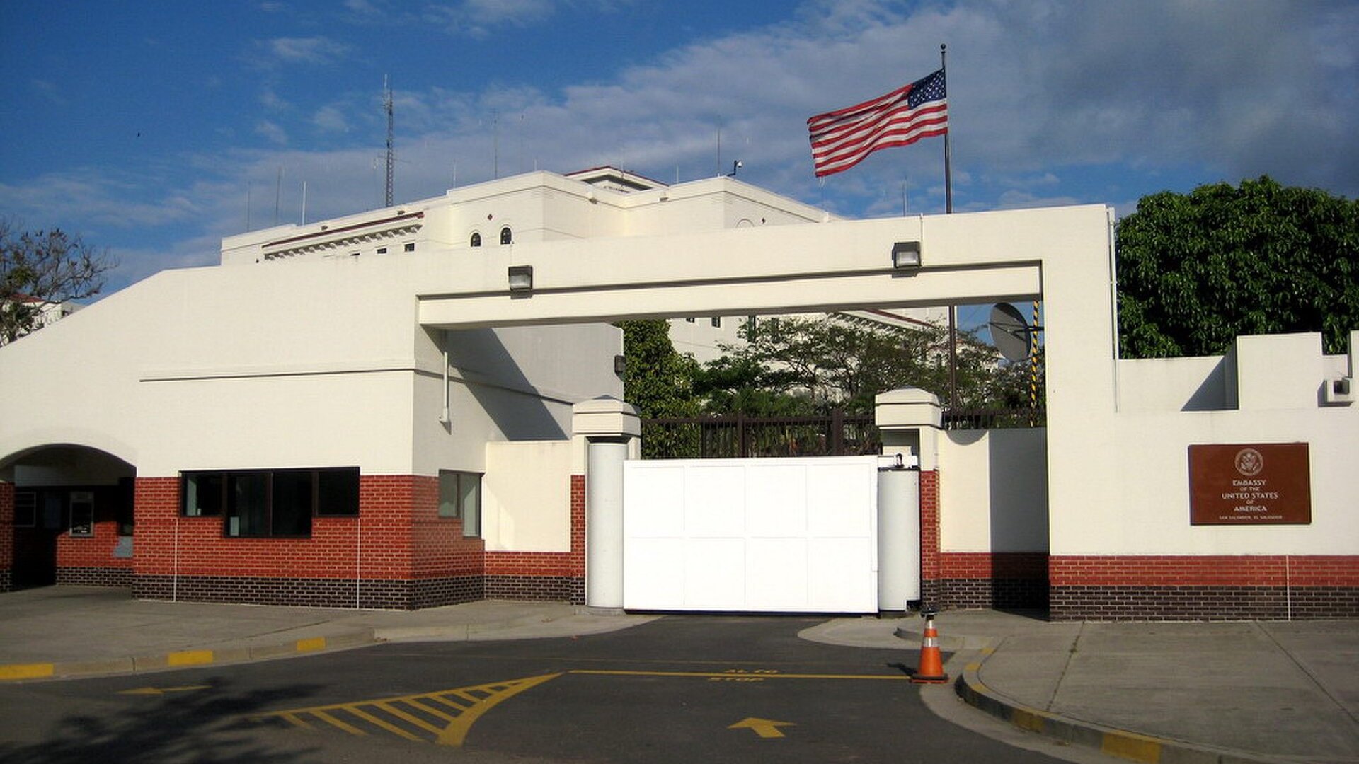 The gate and flag of the United States Embassy in San Salvador, El Salvador, with a white wall, a brick base and a street in front