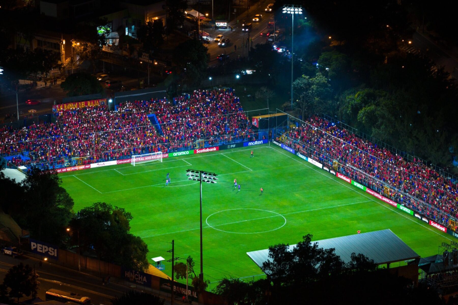 Aerial night view of Guatemala City's El Trébol football stadium under floodlights, its stands packed with fans in red and blue holding up phone lights