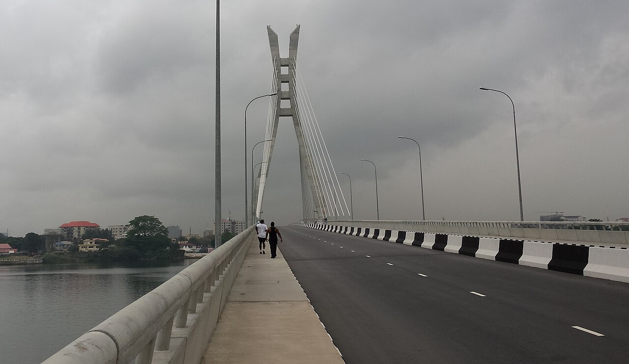 Two pedestrians walk along the Lekki-Ikoyi Link Bridge in Lagos, with its tall cable-stay pylon ahead under a grey sky.