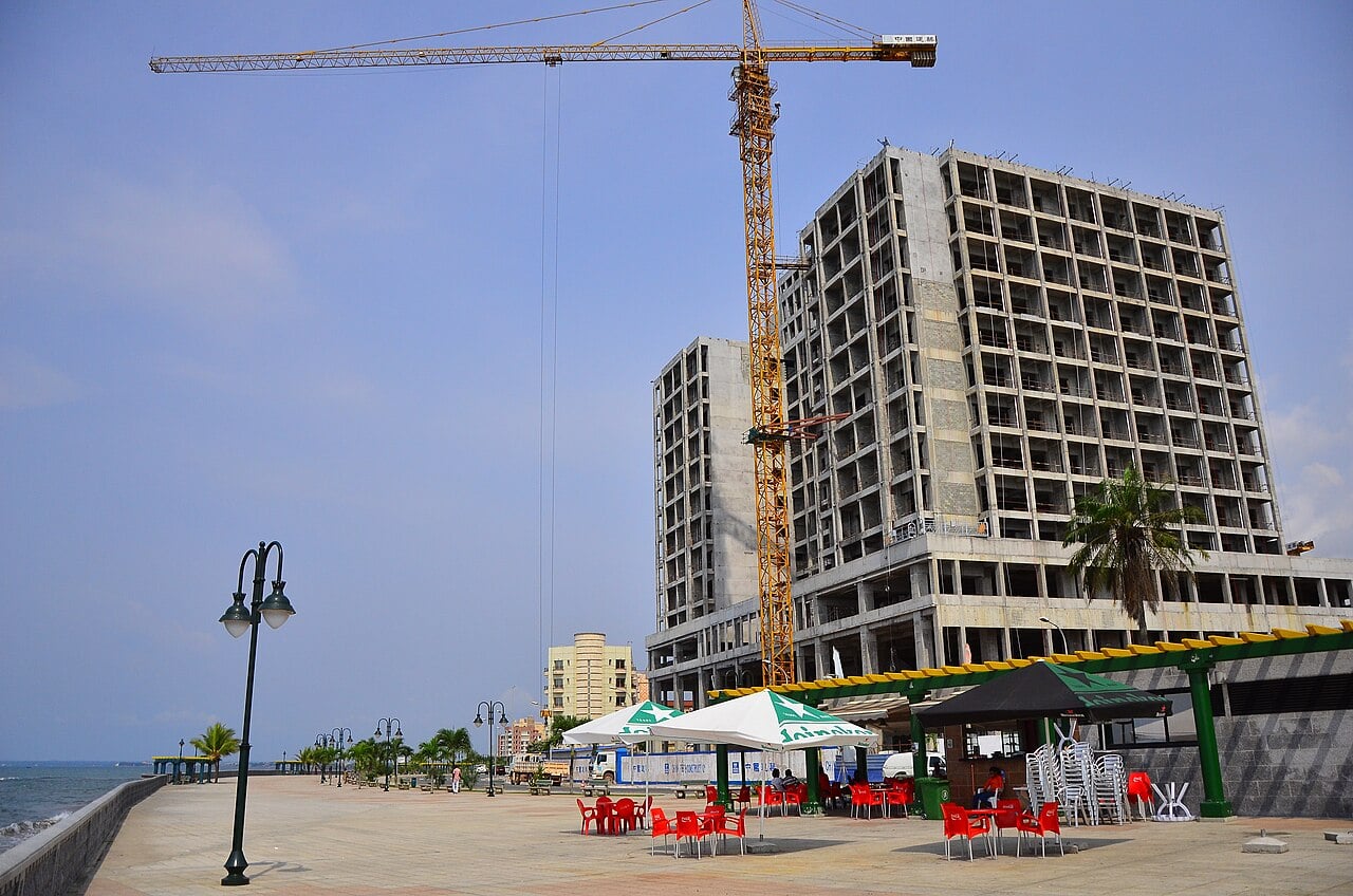 A tower block under construction beside the seafront promenade in Bata, Equatorial Guinea, with a tower crane and a cafe