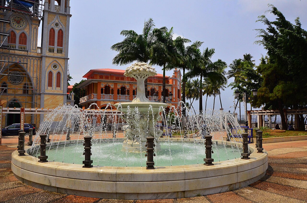 Fountain in a Malabo square with palm trees, an orange colonial-era building and a cathedral tower under scaffolding