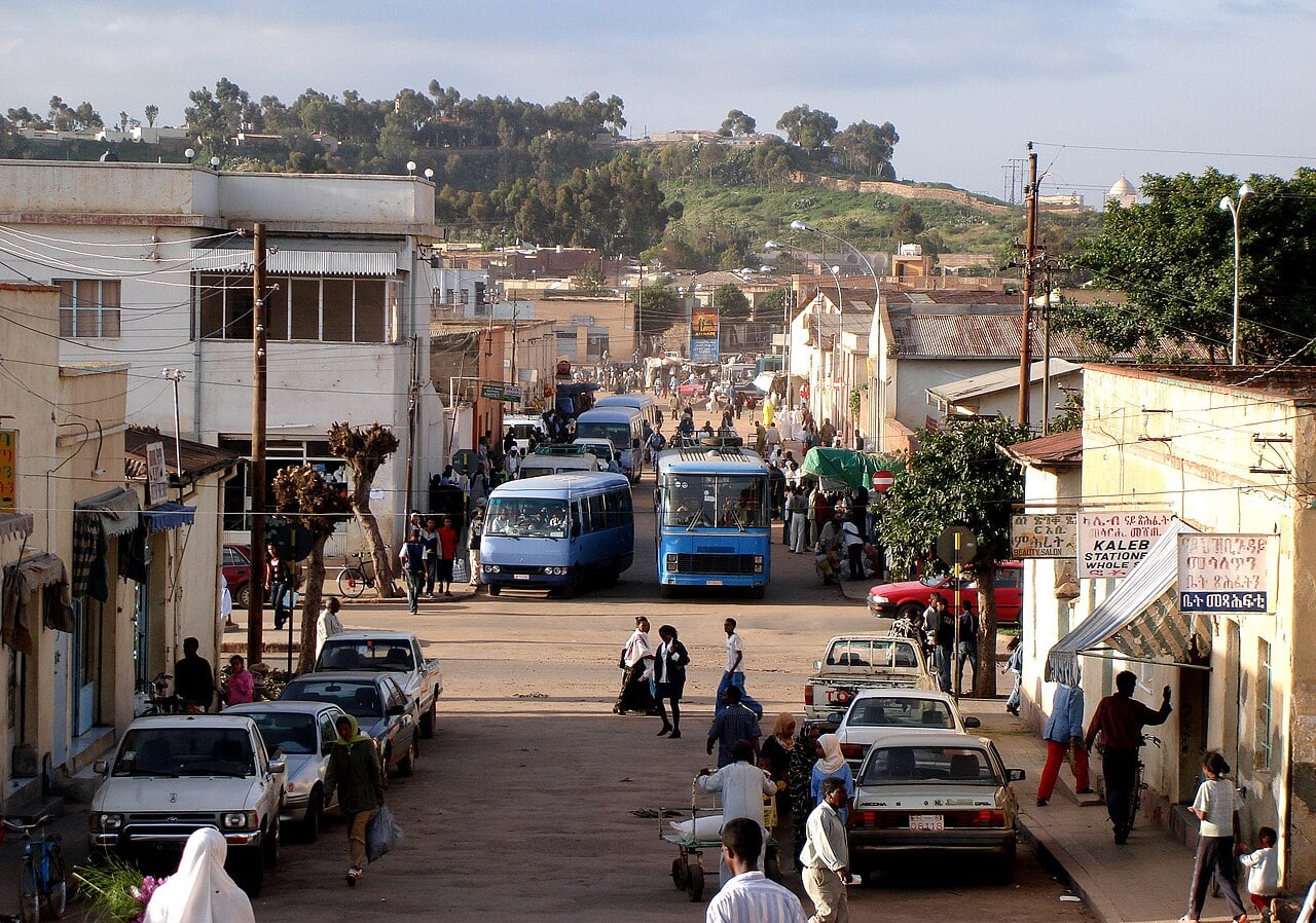 Blue minibuses and pedestrians on a busy street in Asmara, Eritrea, with a green hill behind low buildings