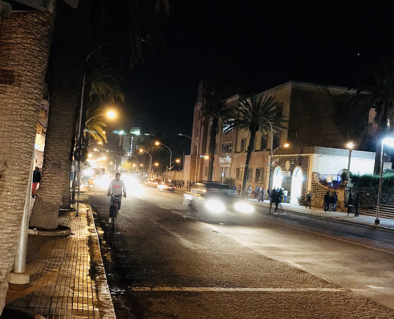 A cyclist and a car on Freedom Avenue in Asmara at night, with palm trees and street lamps beside an Italian-era building