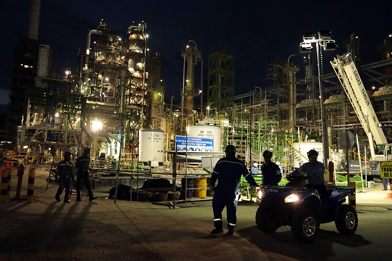 Workers in helmets and overalls and a man on a quad bike in front of lit refinery units and a crane at night