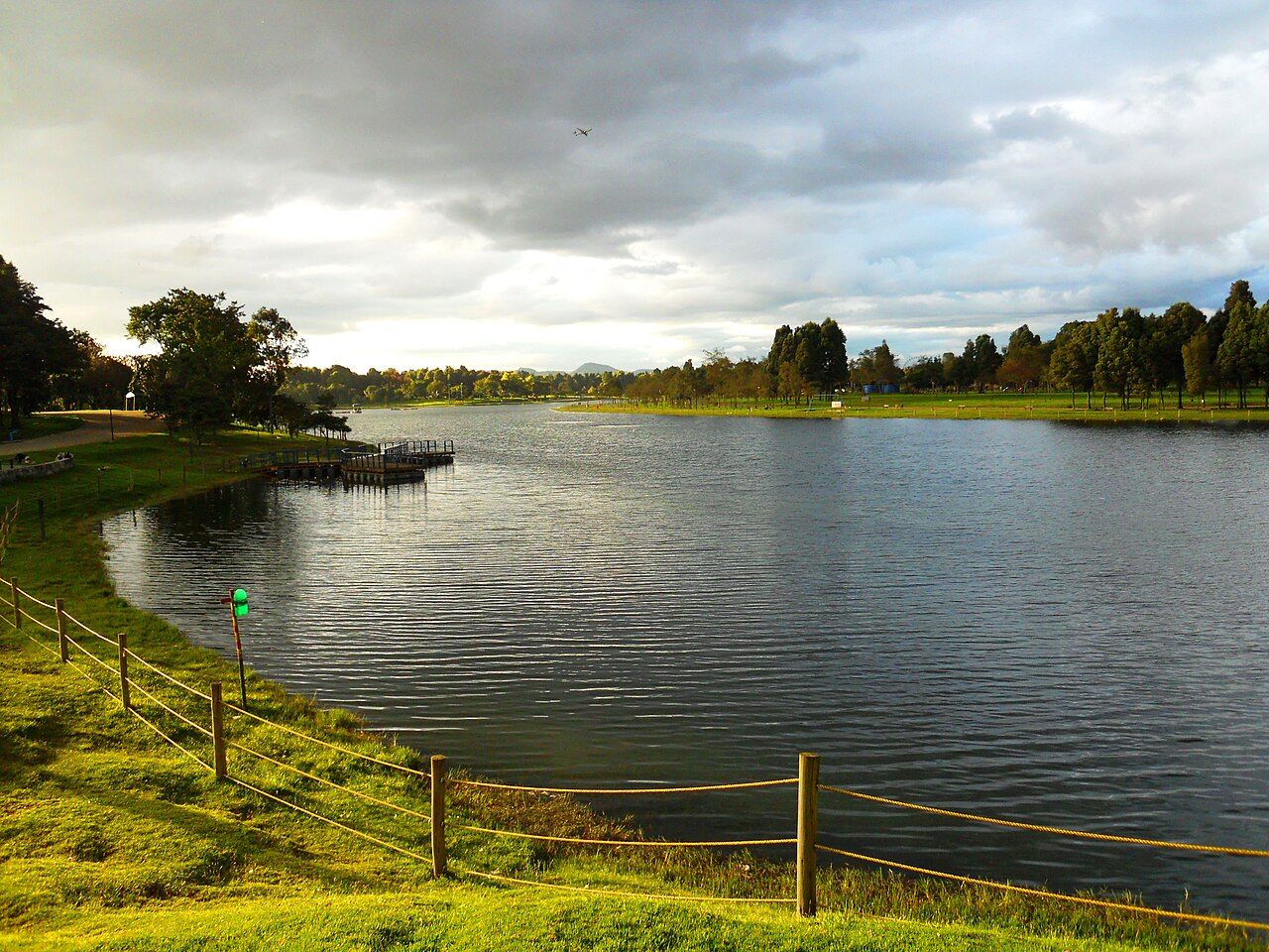 The lake at Parque Simón Bolívar in Bogotá, with a wooden jetty, a rope fence on the grassy bank and trees under a cloudy sky.
