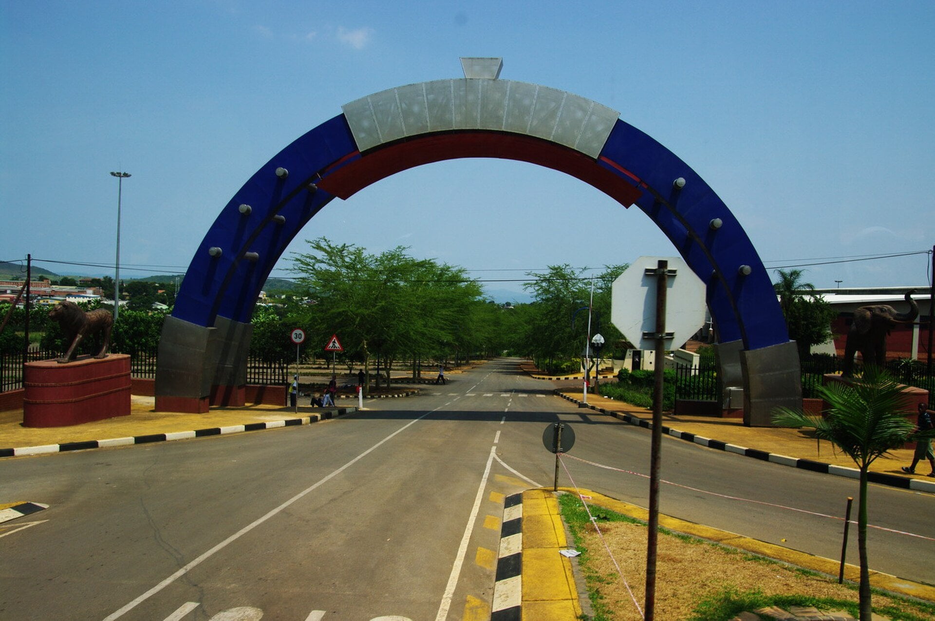A blue entrance arch over the access road to the Mavuso Trade Center in Manzini, Eswatini, with a lion statue on the left