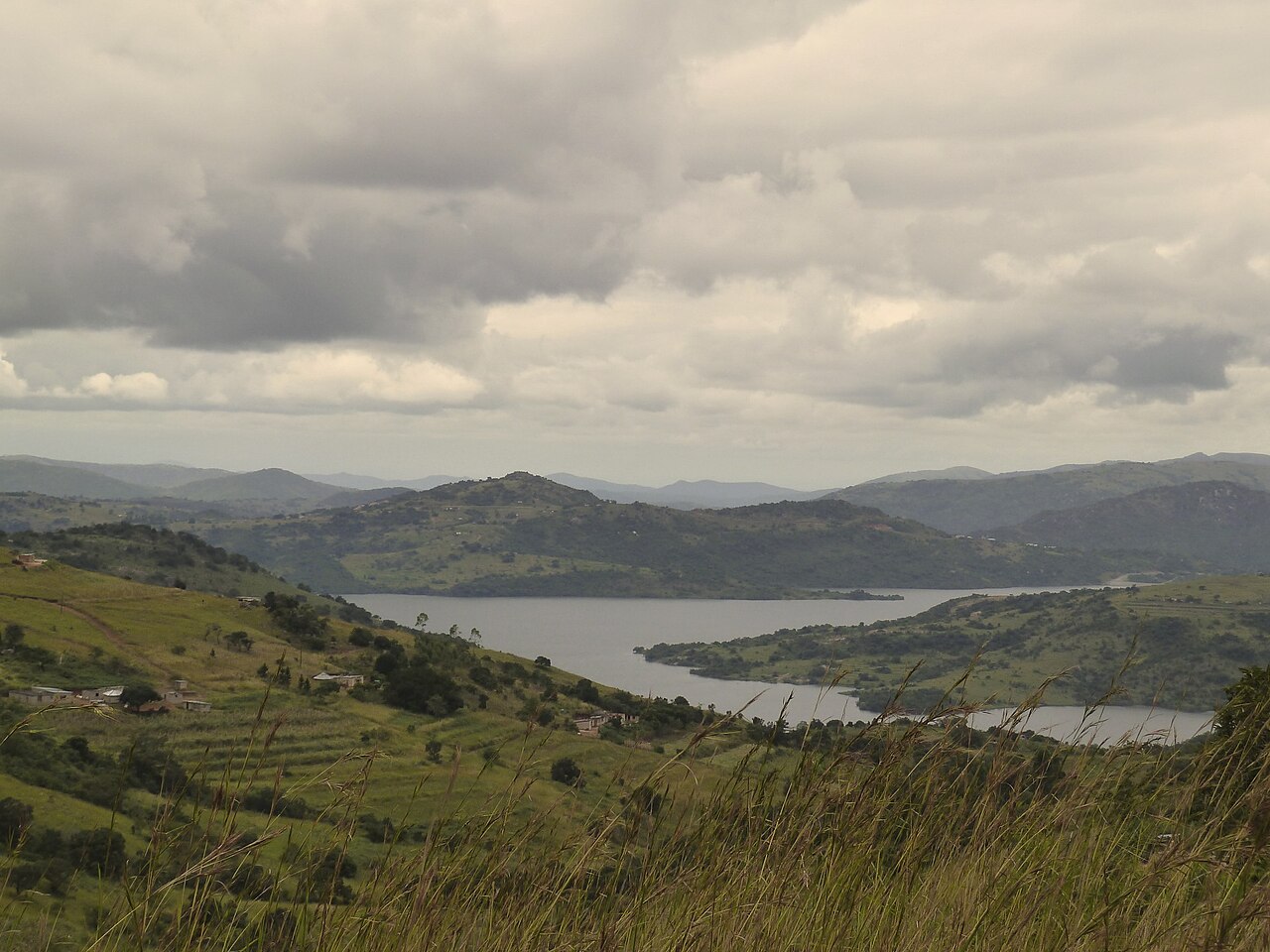 Reservoir of Maguga Dam winding between green hills with scattered homesteads under a cloudy sky