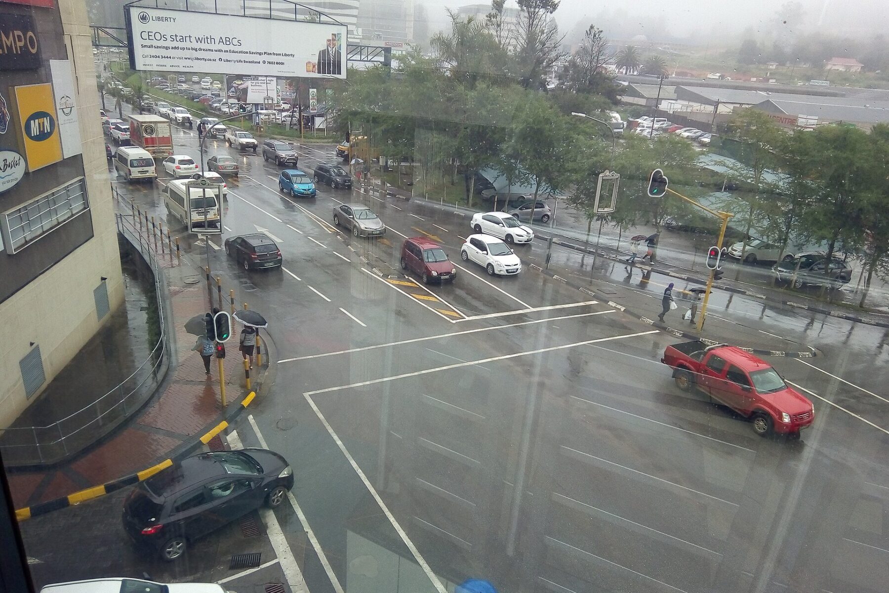 Traffic on a rainy day on Dr Sishayi Road in Mbabane, Eswatini, with an insurance billboard and shop signs