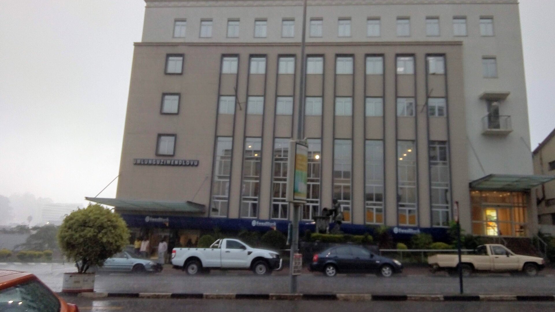 A six-storey cream-coloured Eswatini Bank building in Mbabane on a rainy day, with pickup trucks parked along the street