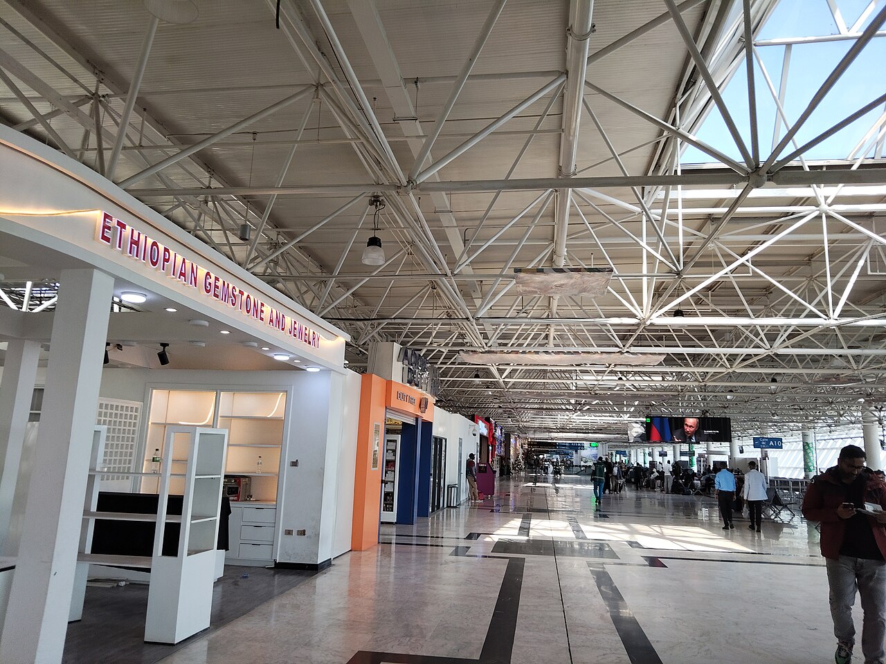 Steel-roofed concourse inside Addis Ababa Bole International Airport with a gemstone shop, duty-free counter and travellers near gate A10.