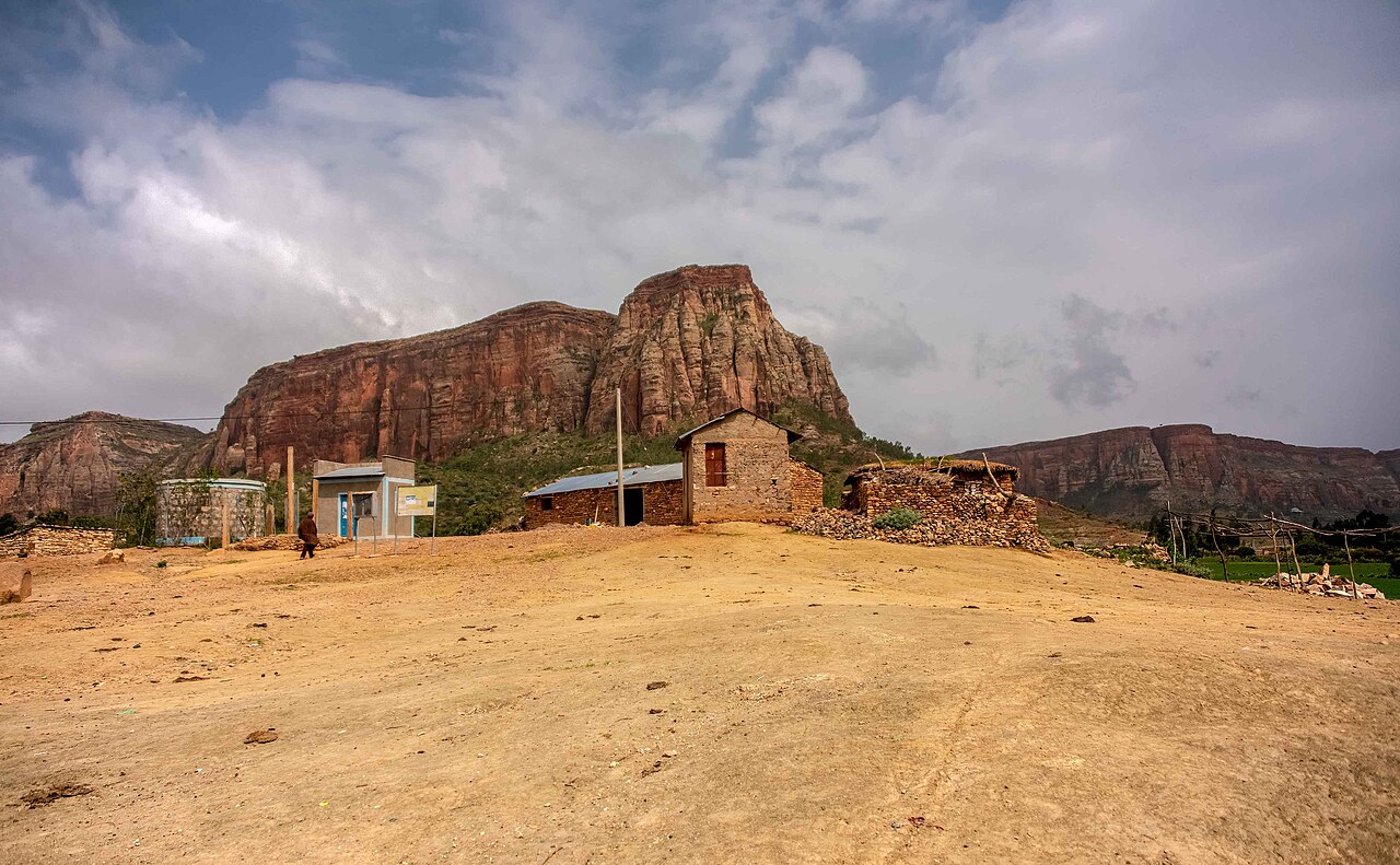 Stone and brick houses on a dusty hilltop in rural Tigray, with tall red sandstone cliffs and cloudy sky behind