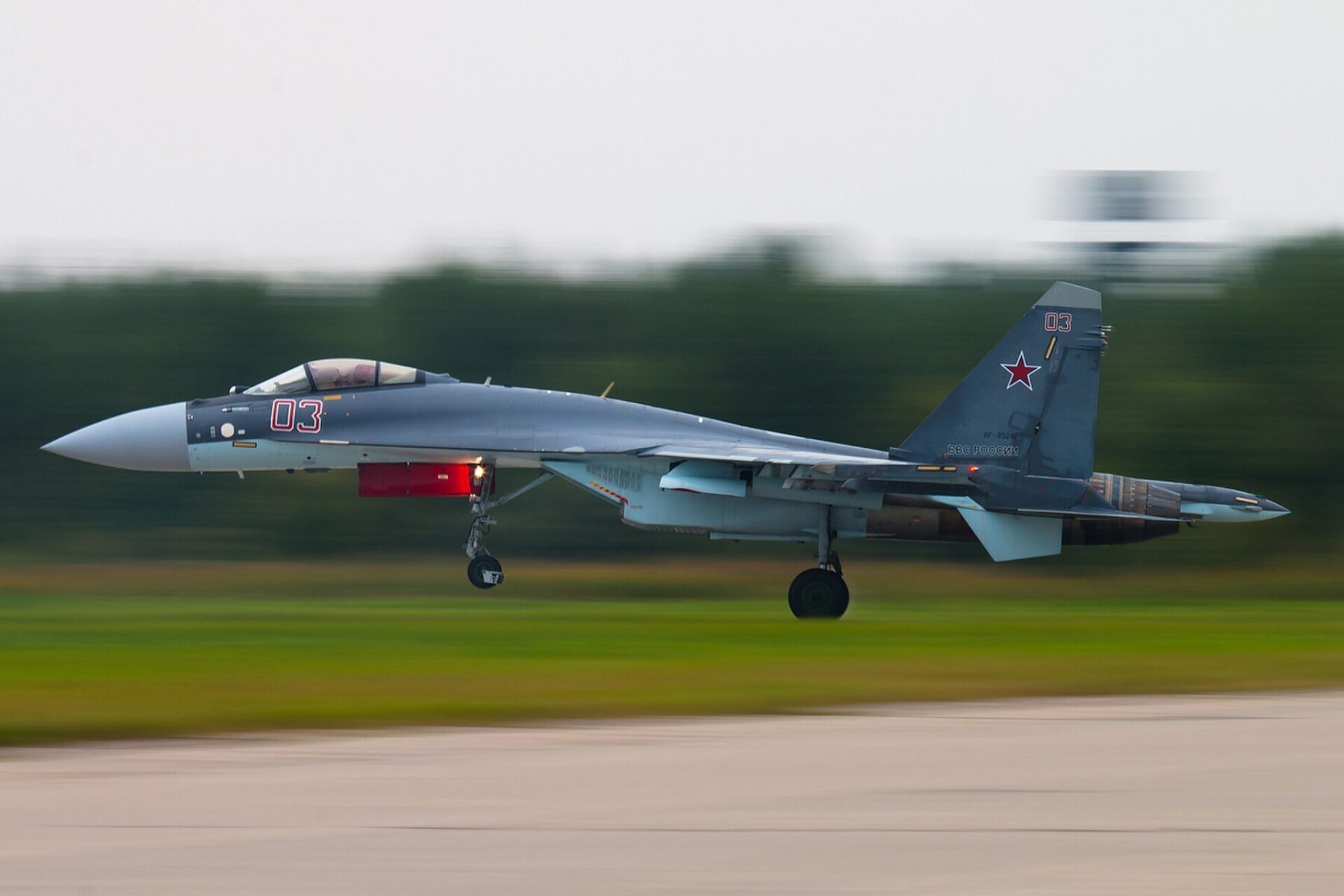 A grey Russian Air Force Su-35 fighter jet lifts off a runway at speed, with a blurred green tree line behind it