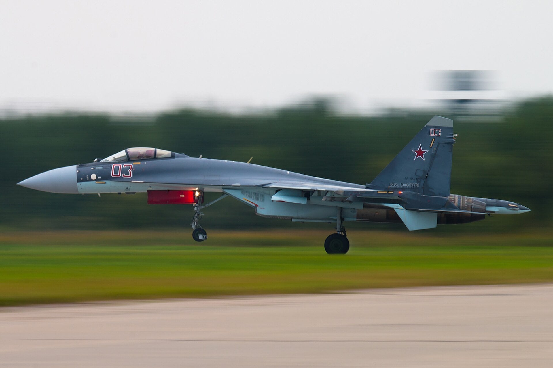 A grey Russian Air Force Su-35 fighter jet lifts off a runway at speed, with a blurred green tree line behind it