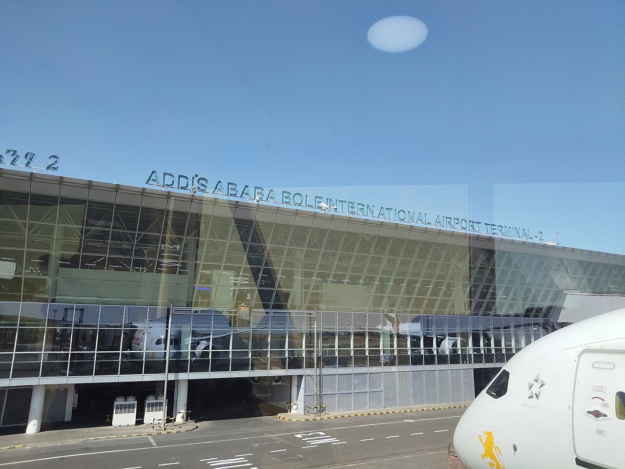 Glass front of Terminal 2 at Addis Ababa Bole International Airport under a blue sky, with an aircraft nose at the right edge