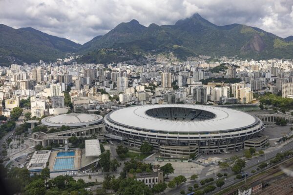 Aerial view of the white-roofed Maracanã stadium in Rio de Janeiro, with apartment blocks and green mountains behind under a cloudy sky