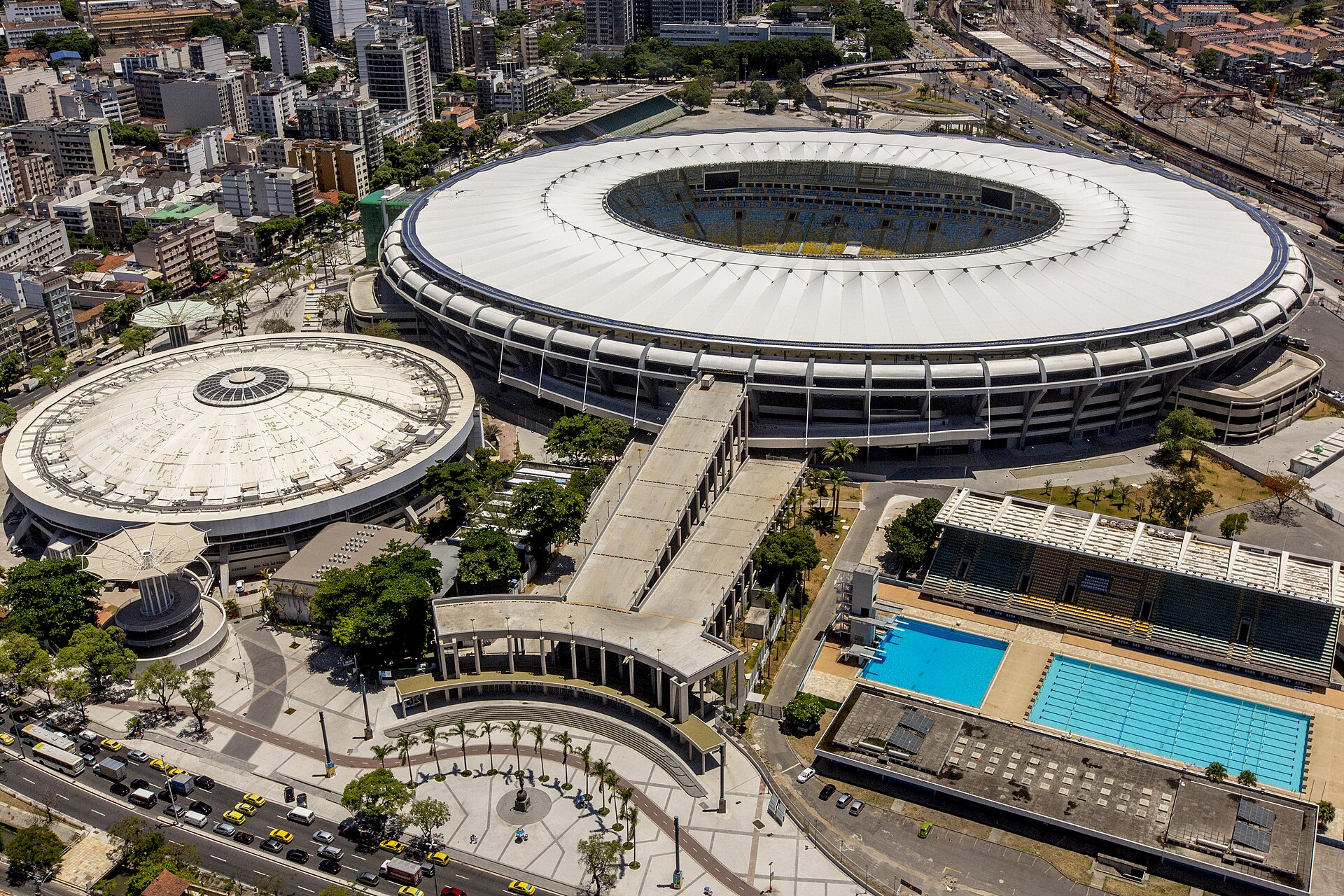 Aerial view of the white oval roof of the Maracanã stadium with the domed Maracanãzinho arena, a long pedestrian ramp and swimming pools beside it
