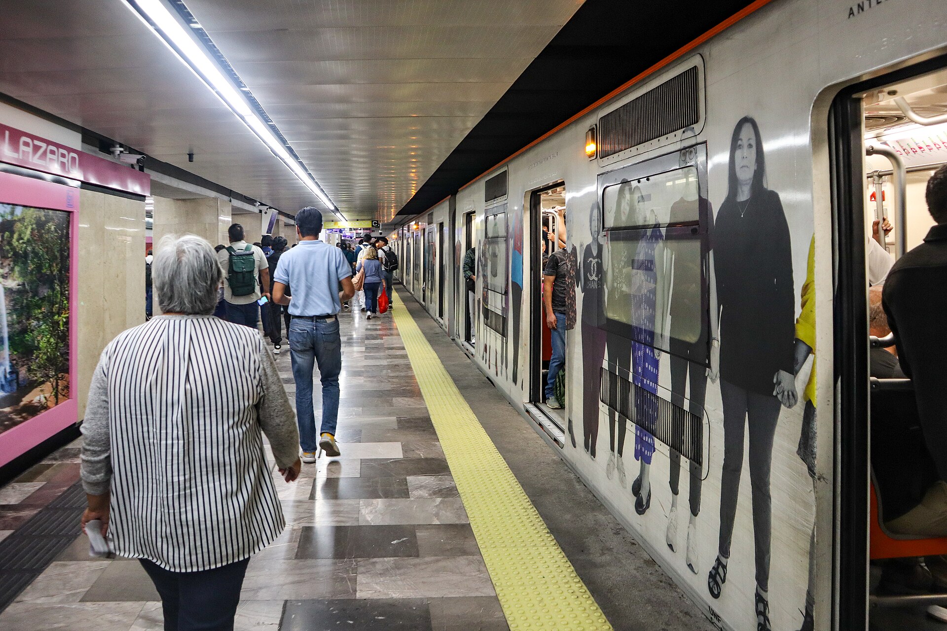 Passengers walking along a Mexico City Metro platform beside a train wrapped in large black-and-white portraits at San Lázaro station