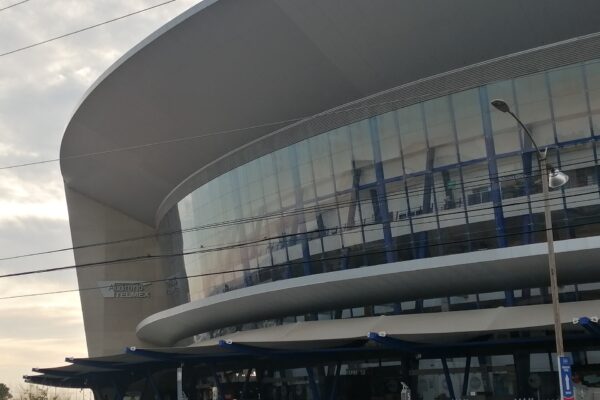 Curved glass and steel facade of the Auditorio Telmex concert hall in Zapopan, with its name sign and a blue entrance canopy