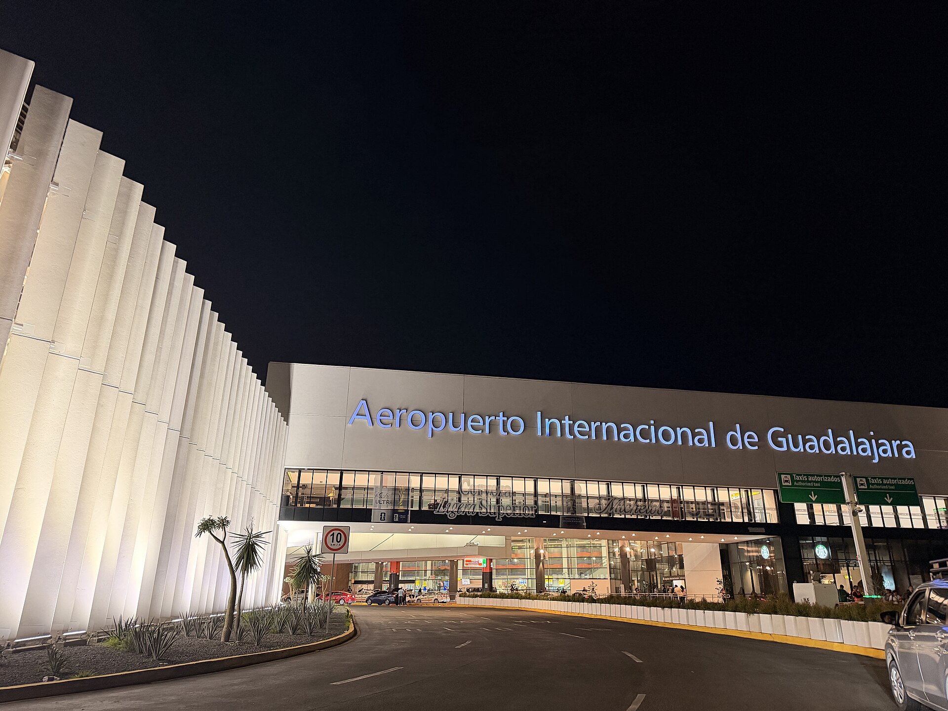 Lit terminal facade with the sign Aeropuerto Internacional de Guadalajara and white vertical fins at night