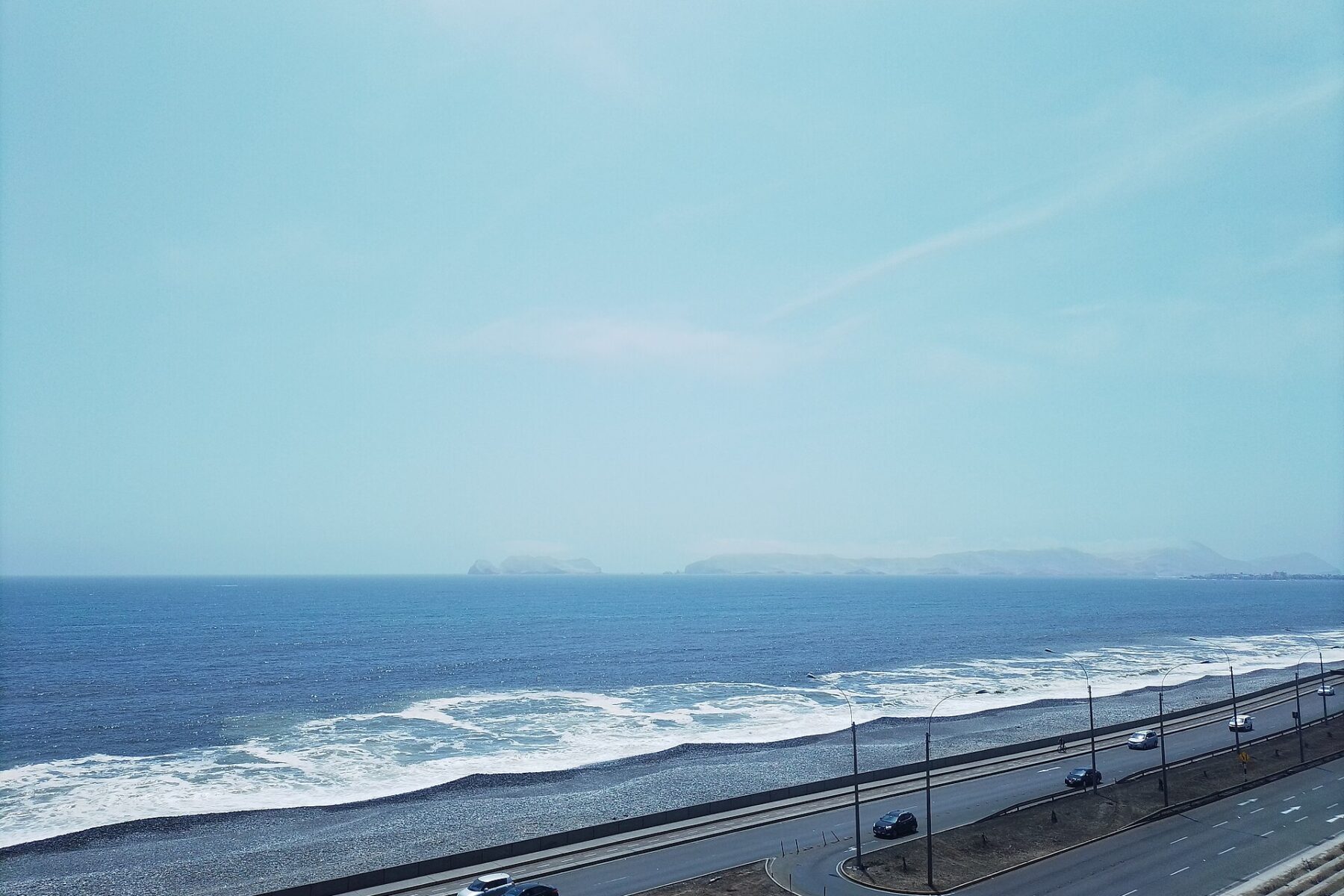 A coastal highway with a few cars runs beside a pebble beach and breaking waves, with islands on the horizon under a pale sky