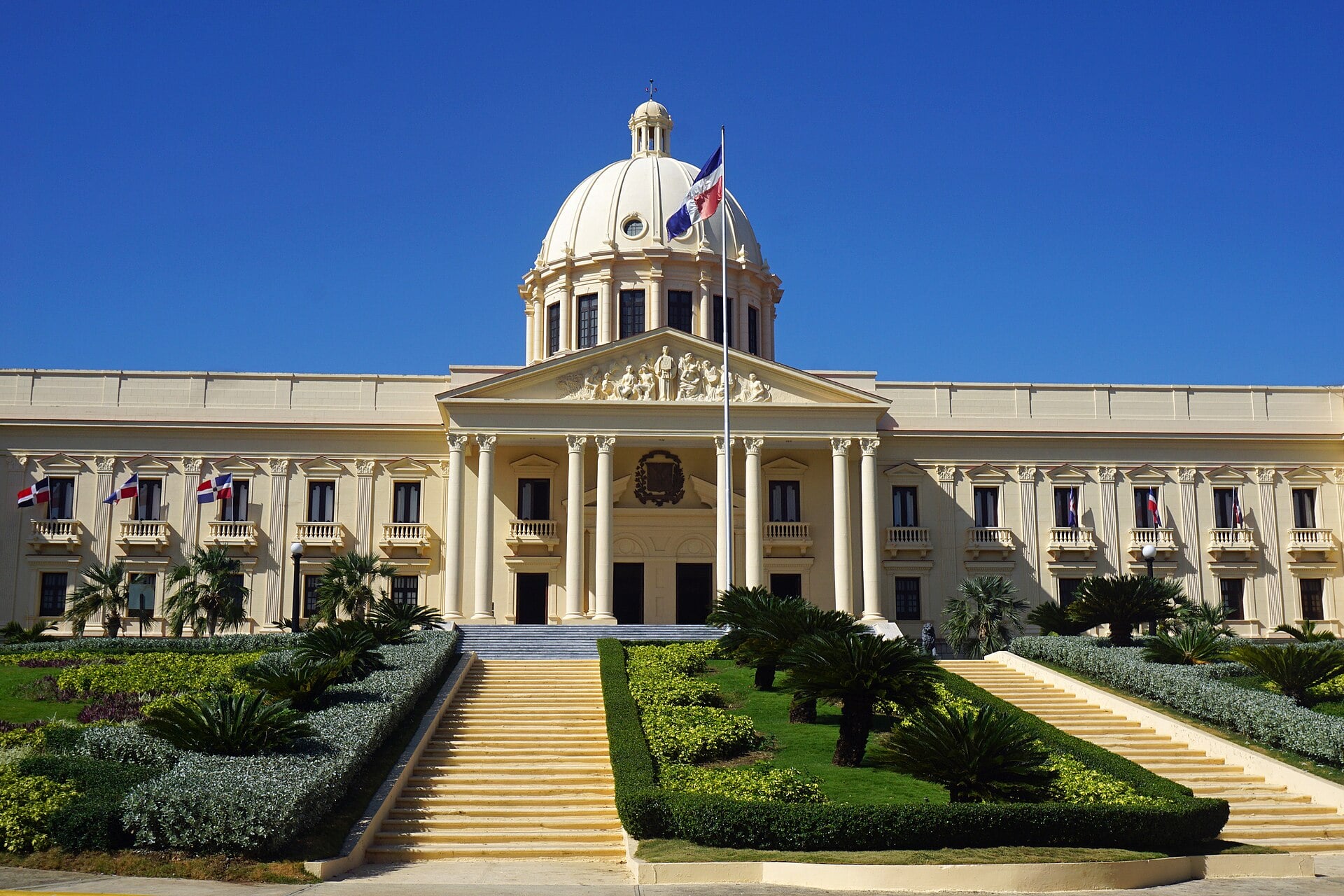 A cream neoclassical palace with a white dome, columned portico and Dominican flags, with stairs and trimmed hedges in front under a deep blue sky