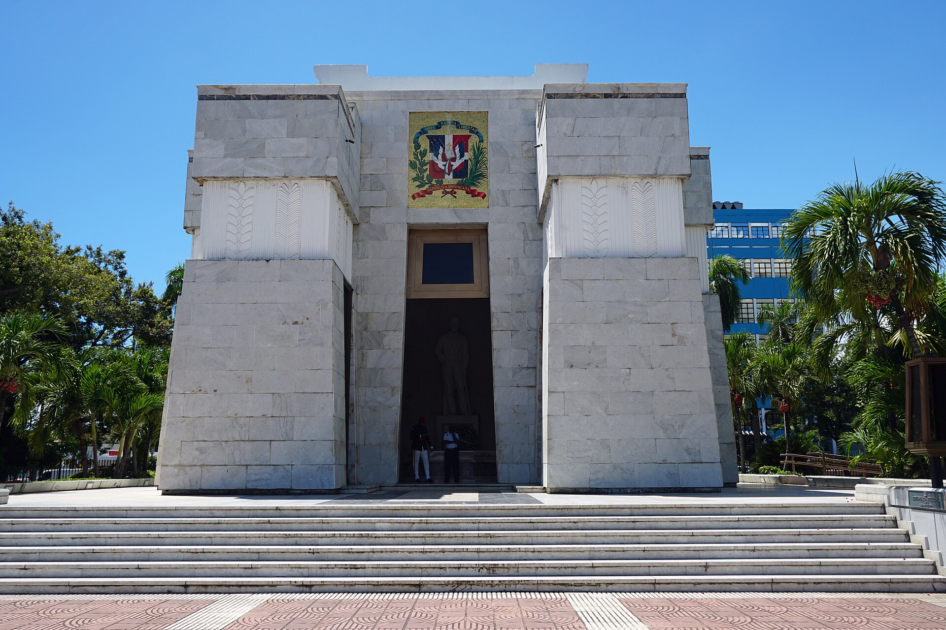 A white marble monument with two massive pillars, the Dominican coat of arms above a dark entrance and wide stone steps in front, under a clear blue sky