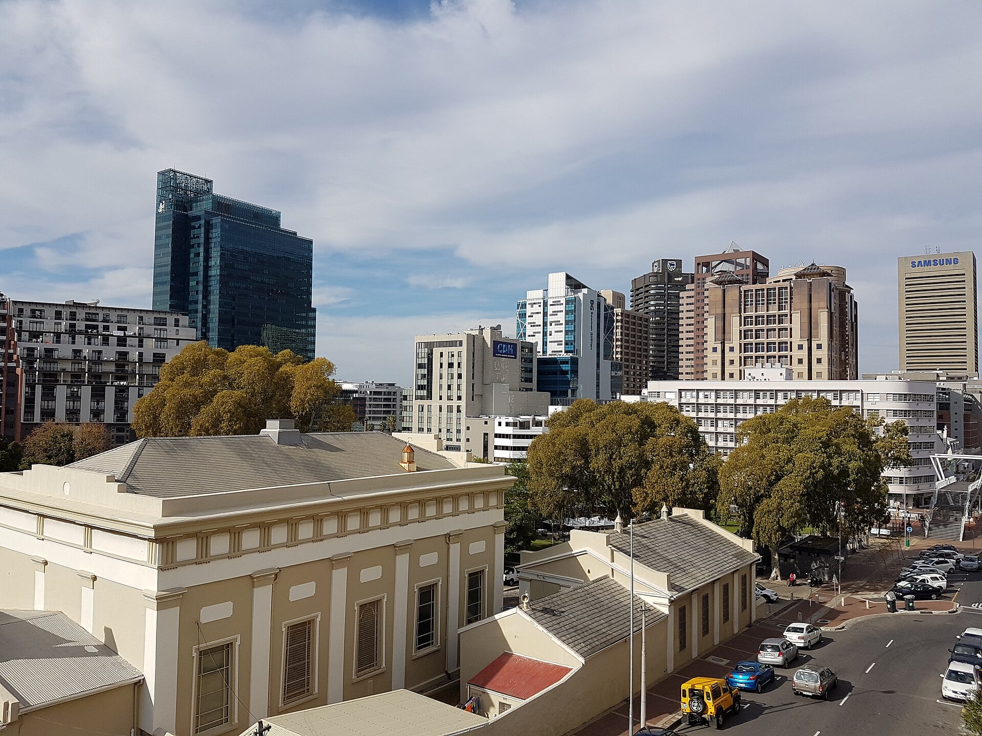 Office towers of central Cape Town rise behind a low cream colonial building and green trees under a cloudy sky