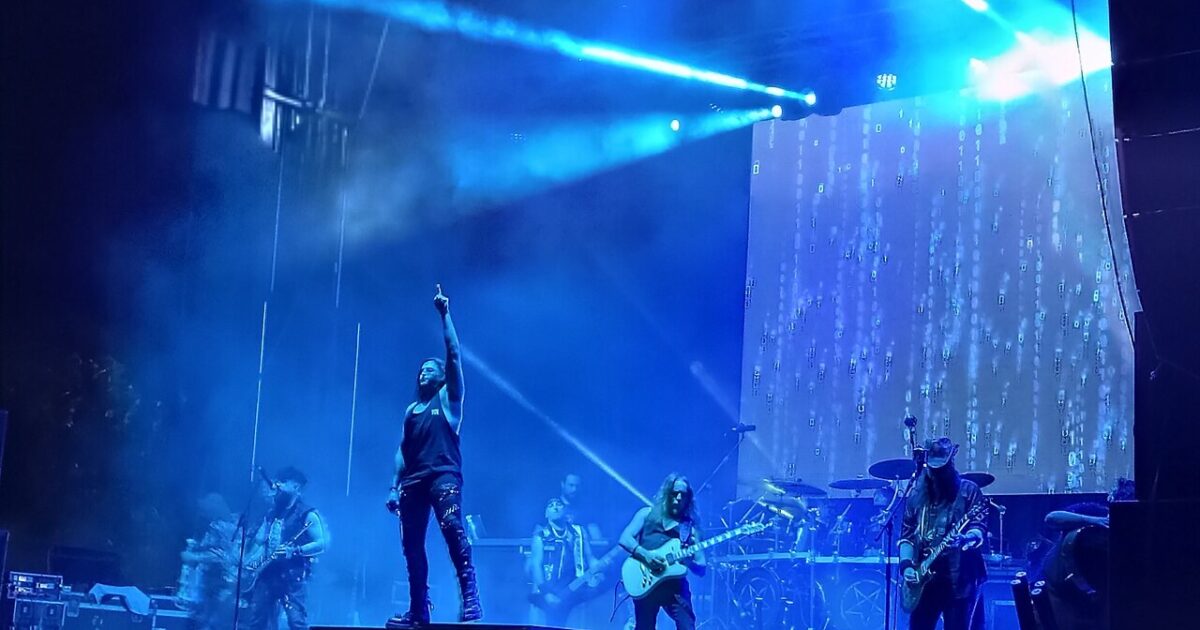 Mägo de Oz on stage under blue lights during a concert in July 2025, the singer pointing upward