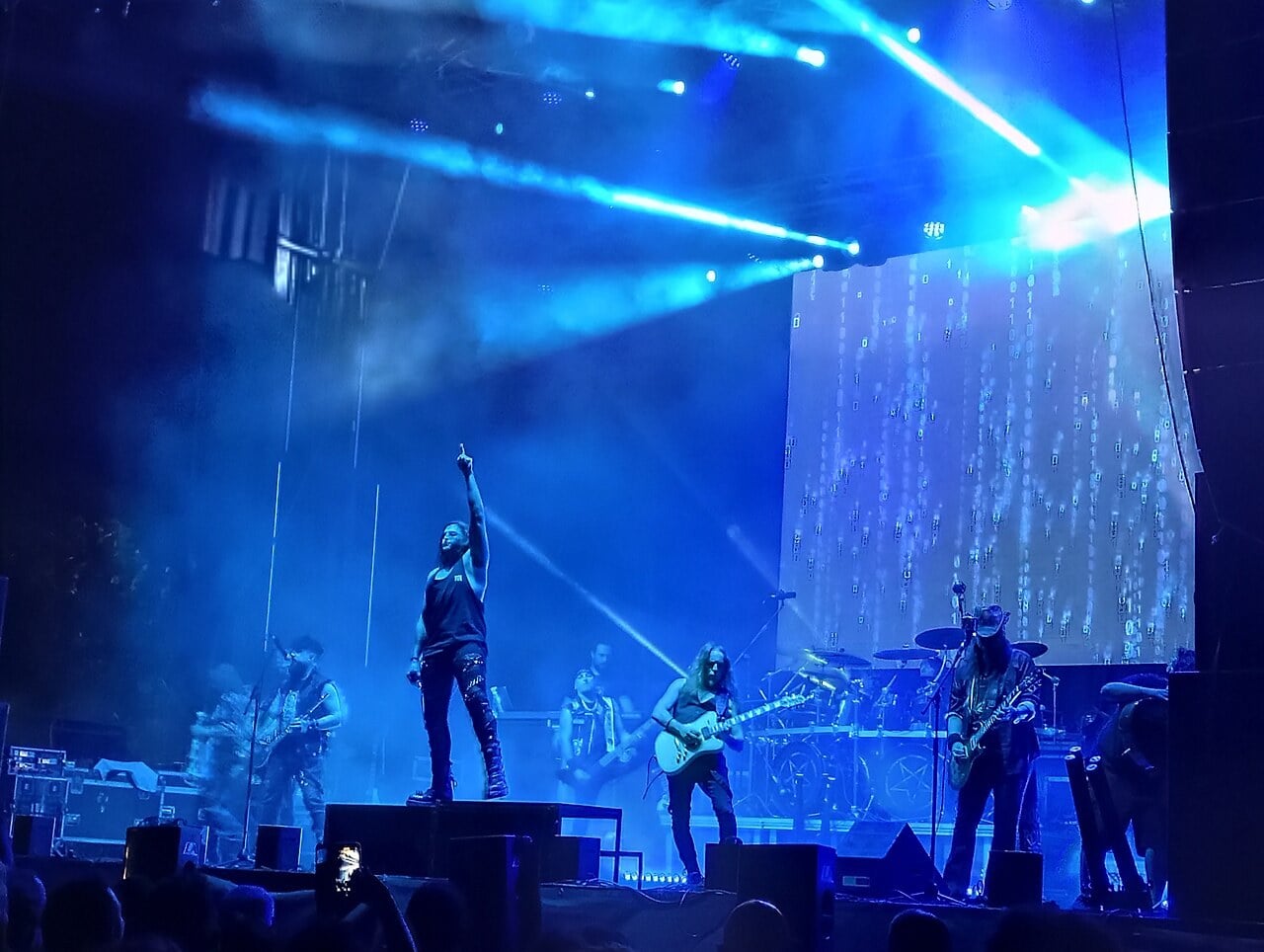 Mägo de Oz on stage under blue lights during a concert in July 2025, the singer pointing upward