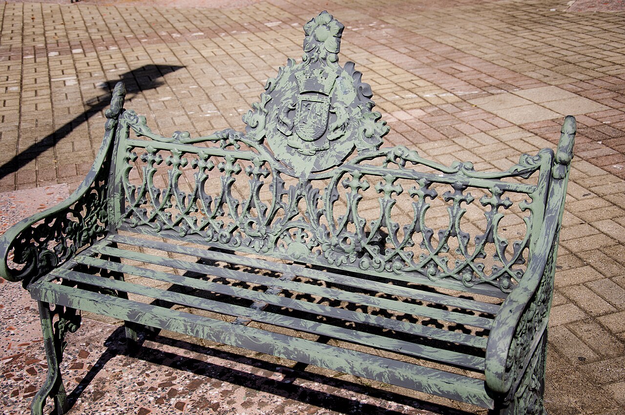 A green wrought-iron bench with an ornate back on a brick-paved public square in Fajardo, Puerto Rico