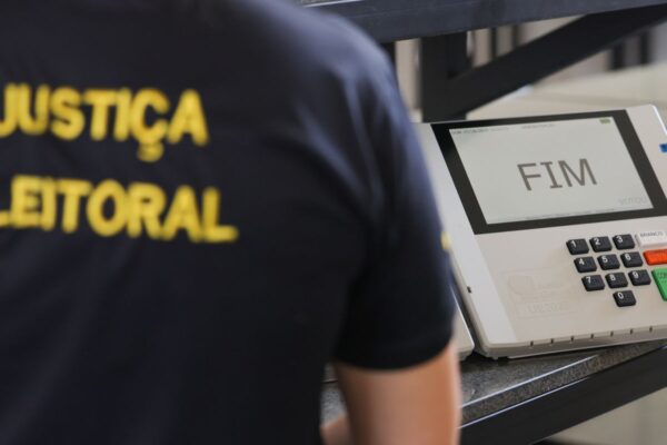 A person in a Justiça Eleitoral shirt stands beside a Brazilian voting machine whose screen reads FIM