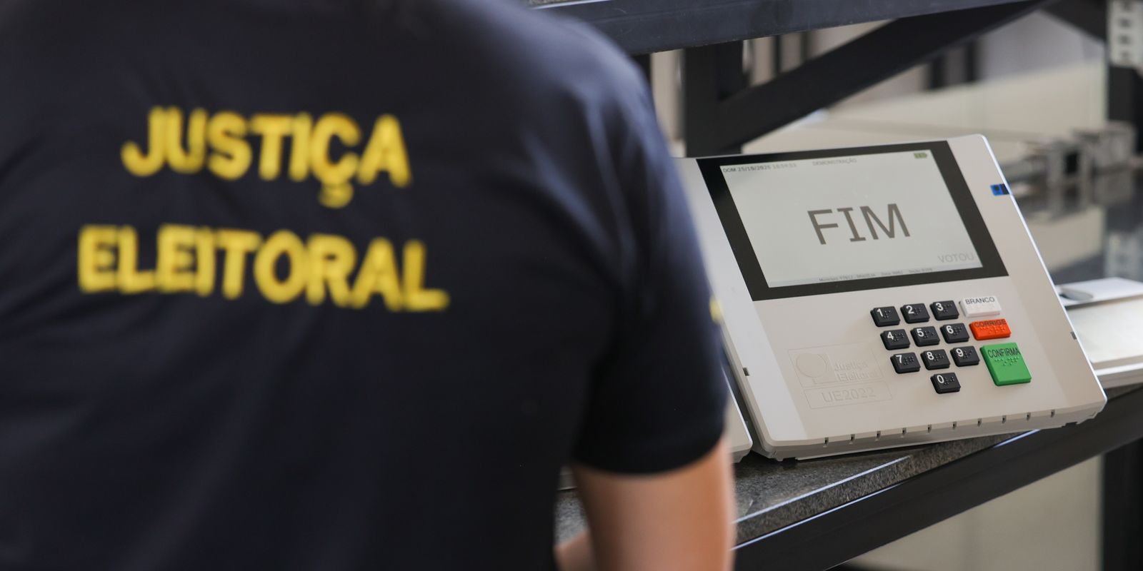 A person in a Justiça Eleitoral shirt stands beside a Brazilian voting machine whose screen reads FIM
