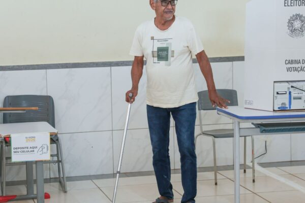 An elderly voter with a walking stick stands beside a polling booth and a voting machine in a Brazilian polling section