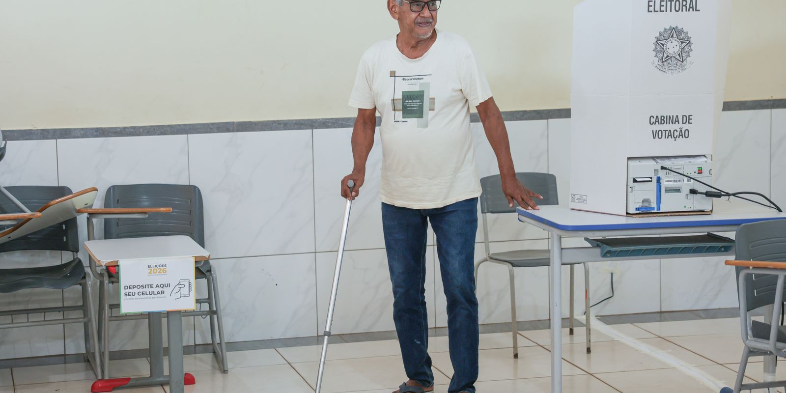 An elderly voter with a walking stick stands beside a polling booth and a voting machine in a Brazilian polling section