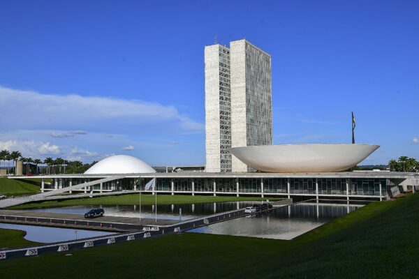 National Congress in Brasília with its twin office towers, the Senate dome at left and the Chamber of Deputies bowl at right, across a reflecting pool under a blue sky