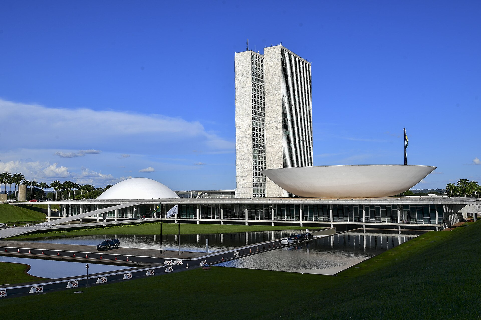 National Congress in Brasília with its twin office towers, the Senate dome at left and the Chamber of Deputies bowl at right, across a reflecting pool under a blue sky