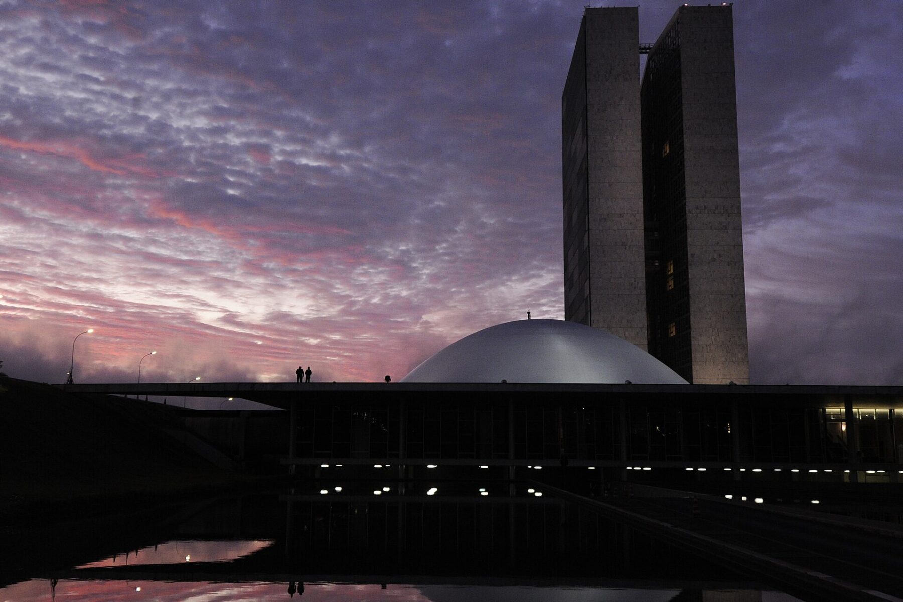 The National Congress in Brasília at dawn, with the twin office towers and the Senate dome silhouetted against a pink and purple sky