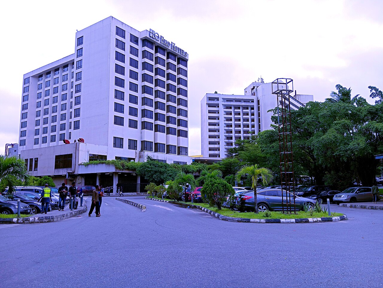 White tower blocks of Eko Hotels & Suites behind a palm-lined forecourt with parked cars and people walking, Victoria Island, Lagos