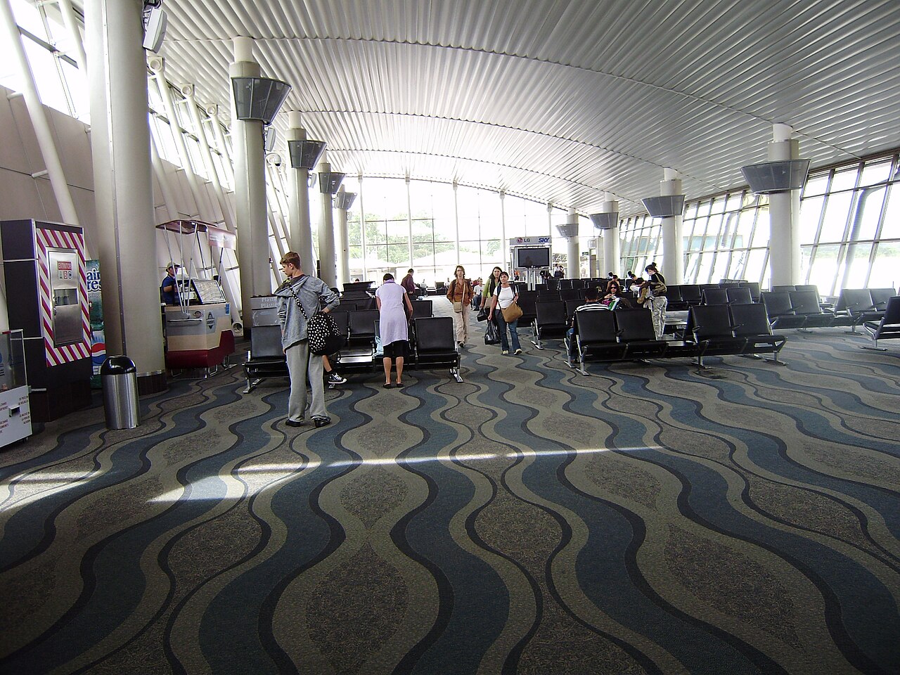 Passengers walking and sitting in a bright waiting hall with a curved metal ceiling at Juan Santamaria International Airport, Costa Rica.
