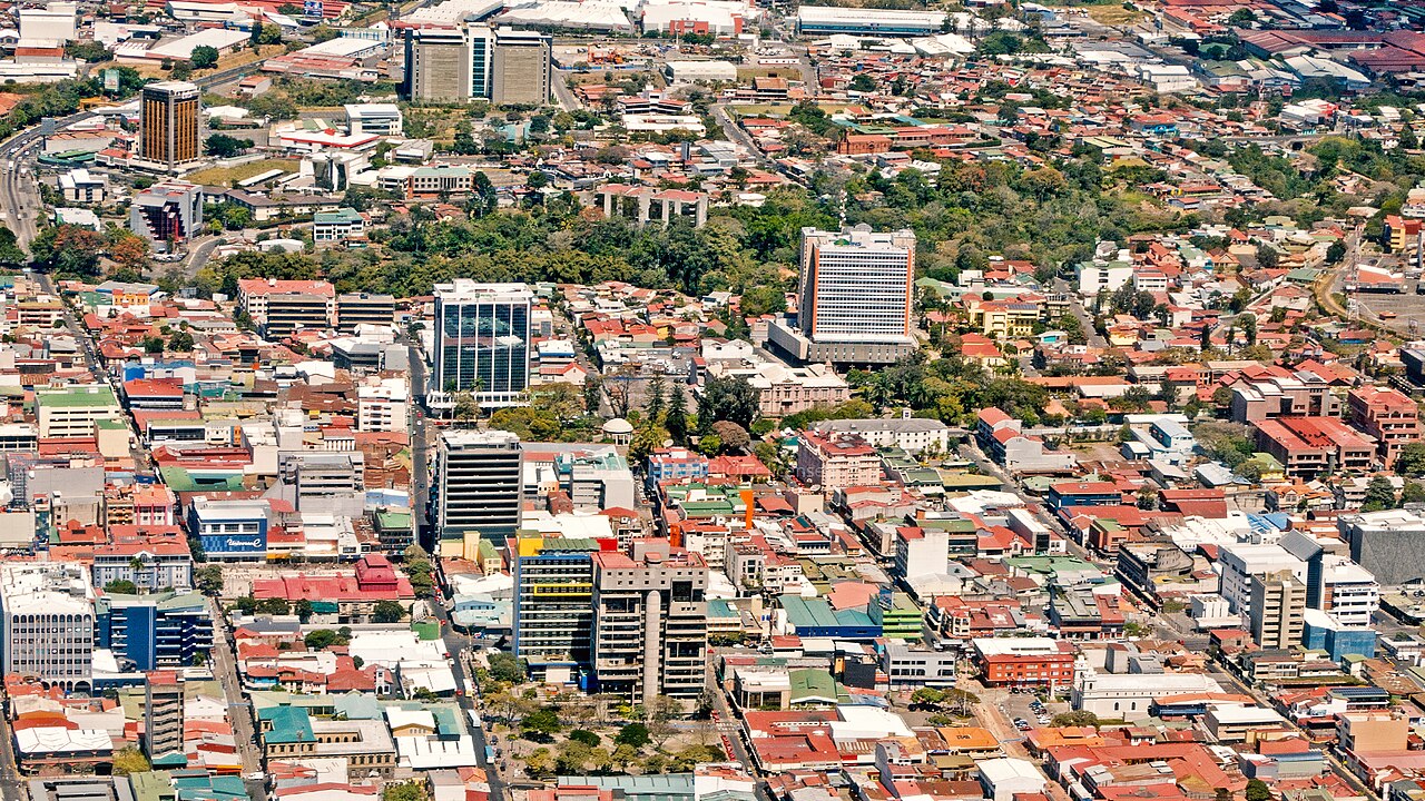 Aerial view of downtown San José, Costa Rica, with office towers and red-roofed low-rise buildings among green parks.