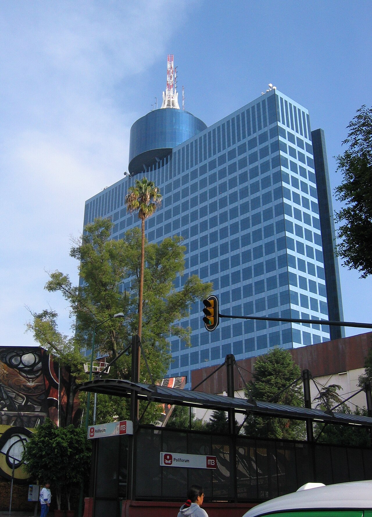 Blue glass World Trade Center tower in Mexico City rising behind a Metrobús station sign that reads Poliforum, with a colourful mural on the left.