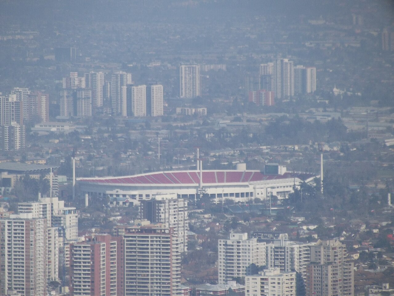 The red-seated Estadio Nacional stadium in Santiago, seen from afar over apartment blocks and the city haze.