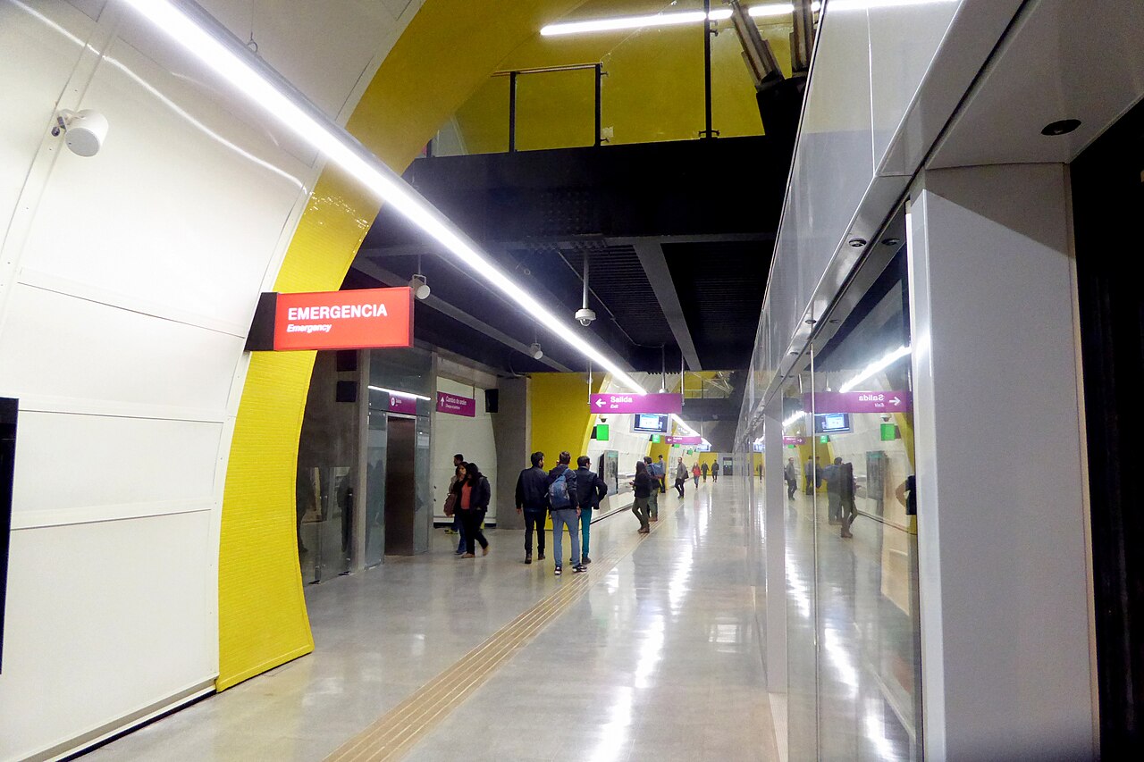 Passengers walk along a Santiago Metro Line 6 platform with yellow arches and purple exit signs.