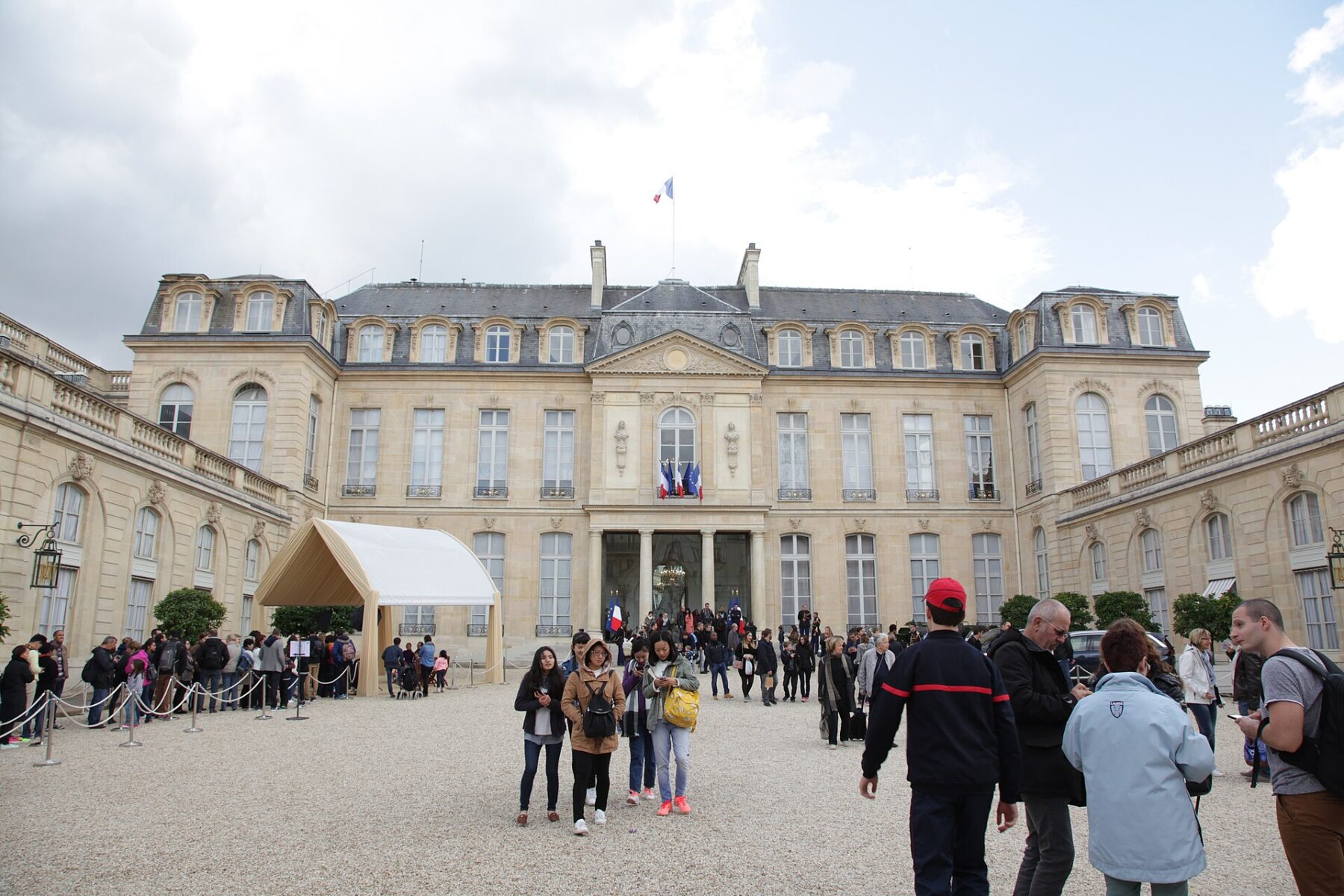 Visitors in the gravel courtyard of the Élysée Palace in Paris, with the French flag above the entrance